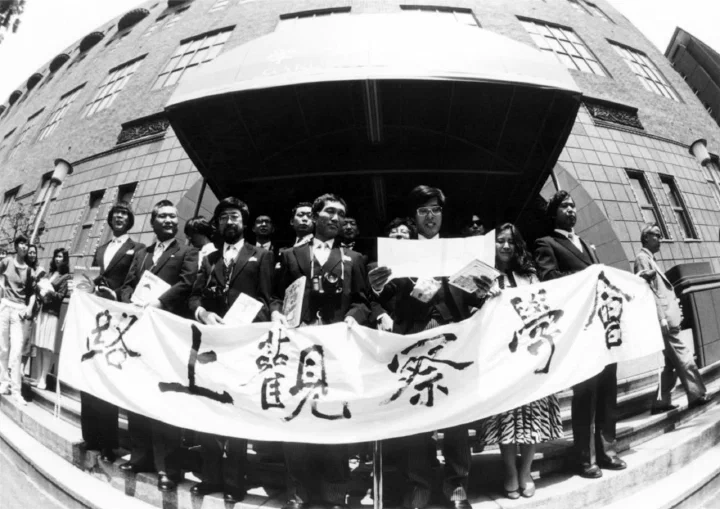 Photographie en noir et blanc de la cérémonie de fondation de la Société d'observation urbaine, le 10 juin 1986. Un groupe de personnes pose devant un bâtiment en tenant une grande banderole calligraphiée en japonais.